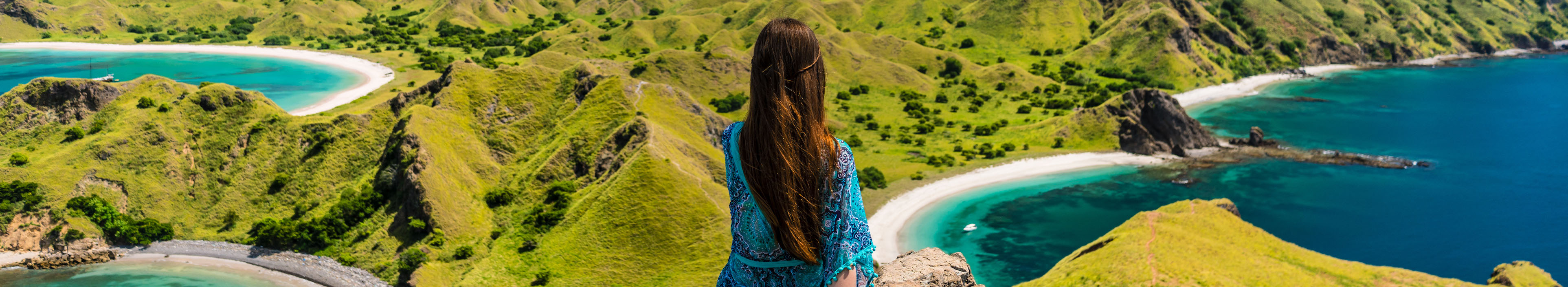 Rückansicht einer jungen Frau, die während der Sommerferien in Indonesien auf dem Gipfel eines Vulkans sitzt und die atemberaubende Aussicht auf die Insel Padar genießt