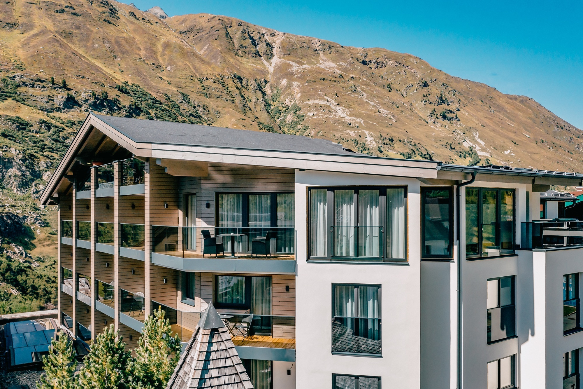 Teilansicht eines modernen Hotelgebäudes mit einer Kombination aus heller Putzfassade und Holzverkleidungen. Die Balkone verfügen über Glasgeländer und bieten einen direkten Blick auf die dahinterliegende, karge Gebirgslandschaft unter strahlend blauem Himmel.