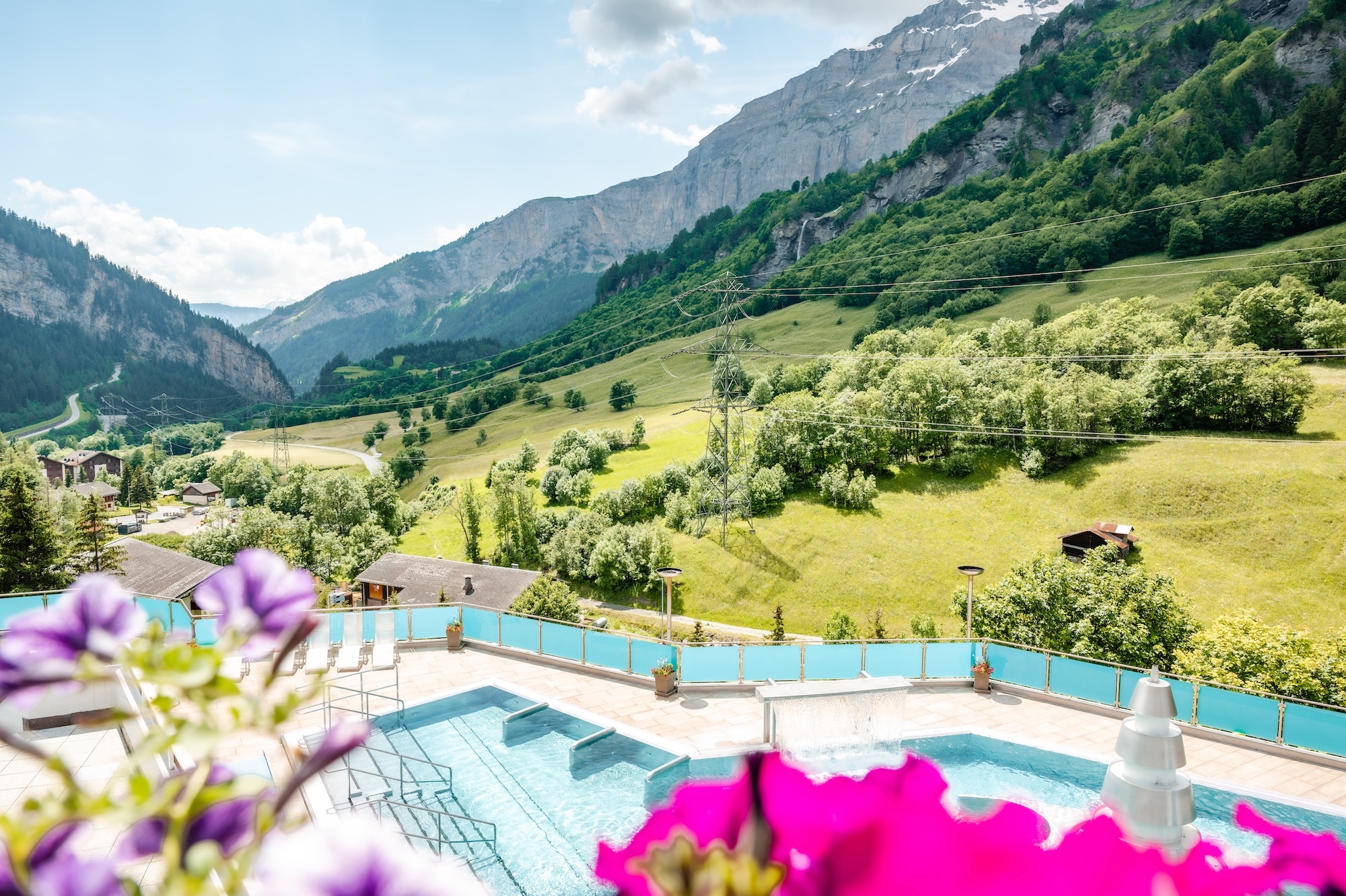 Blick von einer Terrasse mit lila Blumen im Vordergrund auf ein Thermal-Außenbecken. Die Anlage liegt eingebettet in ein grünes Tal mit bewaldeten Hängen und einer steilen Felswand im Hintergrund. Ein Wasserfall stürzt an der gegenüberliegenden Bergseite in die Tiefe.