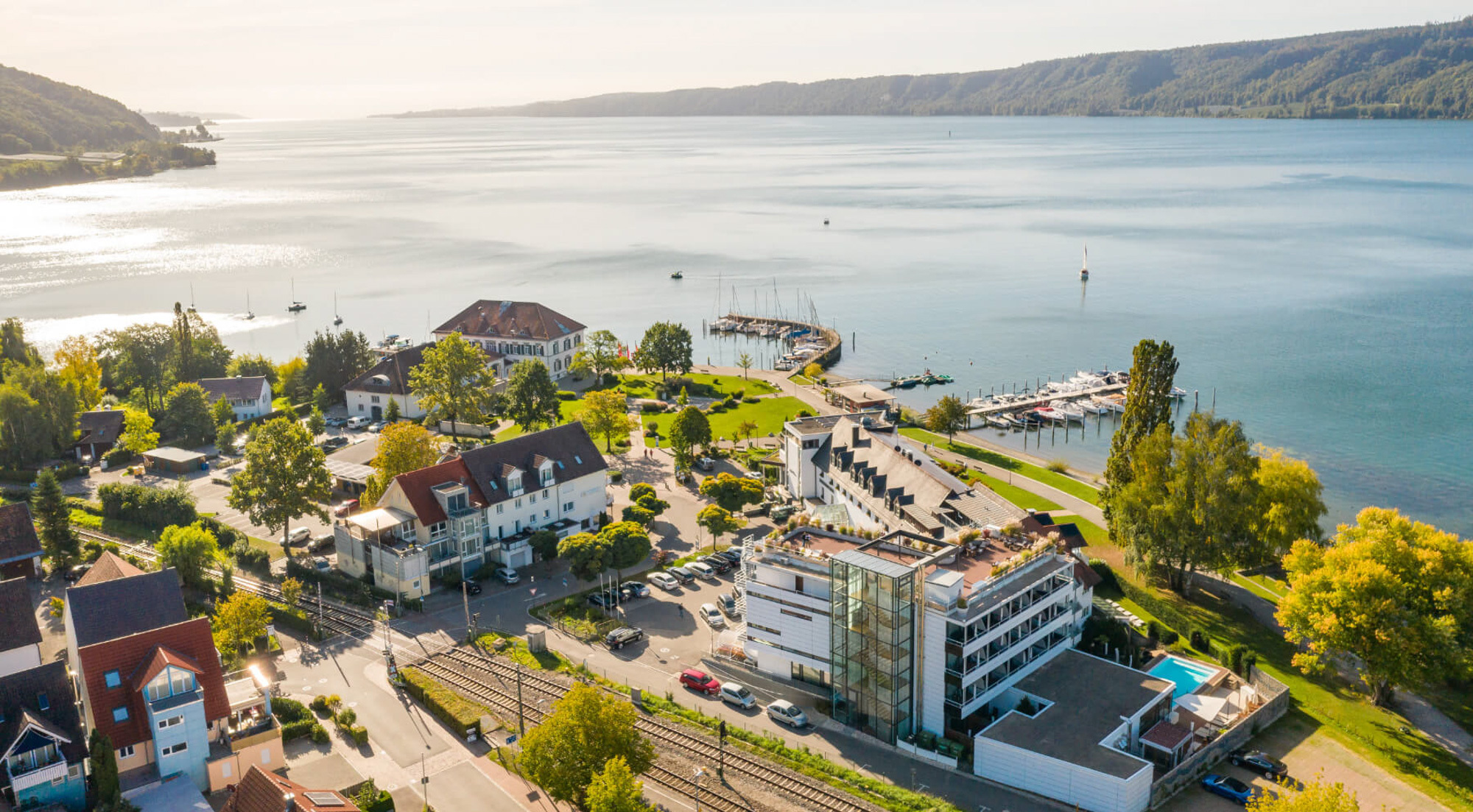 Eine Luftaufnahme einer Hotelanlage direkt am Ufer eines großen, ruhigen Sees. Das Hotel besteht aus mehreren Gebäuden, darunter ein moderner weißer Trakt mit Glasfassade und ein traditionelleres Gebäude mit rotem Dach. Im Hintergrund erstrecken sich bewaldete Hügel entlang des Sees; am Ufer befinden sich kleine Anlegestellen für Segelboote.