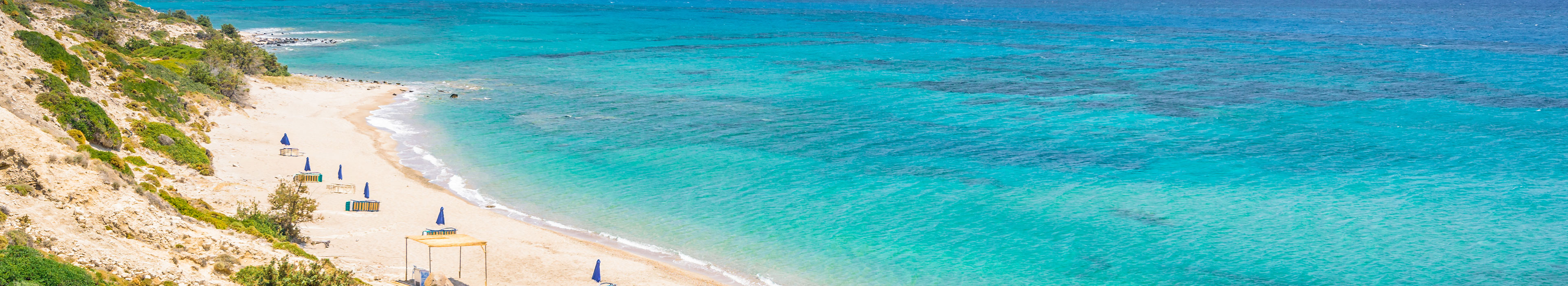 Ein Strand auf Kos mit Sonnenschirmen an der griechischen Ägäis mit türkisfarbenem Wasser und einer malerischen Bucht und Inseln im Hintergrund.