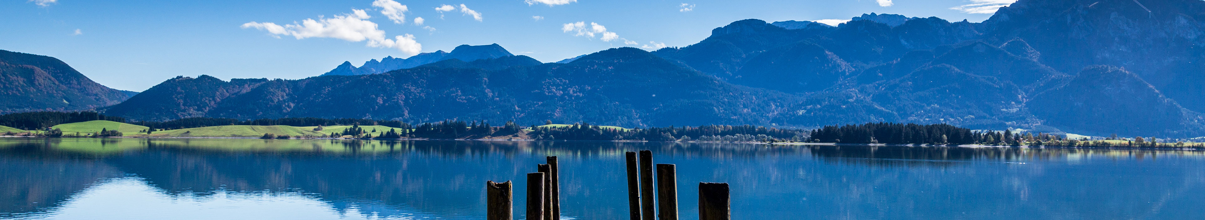 Ein See in Bayern, im Vordergrund ein Steg, der in den See führt, im Hintergrund die Berge. 