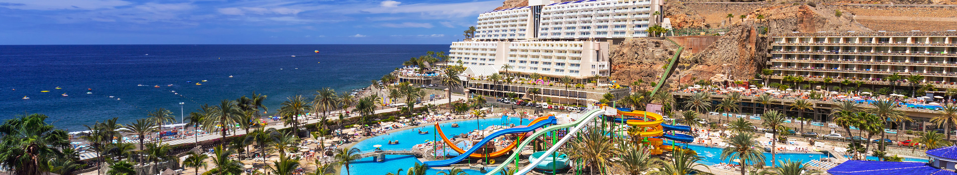 Hotels auf Gran Canaria, im Hintergrund das Meer. Im Vordergrund ein Hotelpool, Palmen, Liegen und Sonnenschirme.